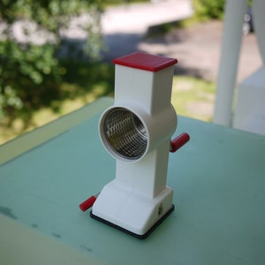 Puede incluir: Un rallador de queso manual blanco y rojo con una superficie de rallado de acero inoxidable. El rallador tiene una base rectangular y un cuerpo cilíndrico con un mango rojo. La parte superior es roja y texturizada.