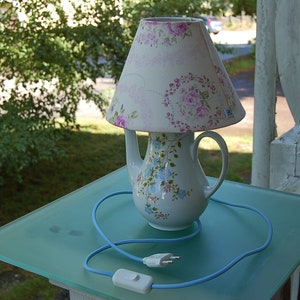 Può includere: Una lampada unica realizzata con una teiera bianca con un design floreale, sormontata da un paralume abbinato. Il paralume e la teiera presentano motivi floreali rosa e blu. Un cavo blu con interruttore e spina completa il design.