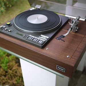 Może przedstawiać: Zabytkowy gramofon TEAC z wykończeniem z ciemnego drewna. Gramofon posiada czarny talerz, srebrne ramię i panel sterowania. Urządzenie jest zamontowane na białym cokole ze szklaną półką.