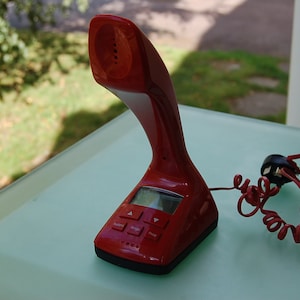 May include: A red, retro-style telephone with a curved design. The phone has a small screen and several buttons. A red, coiled cord extends from the base. The phone is on a light green surface.