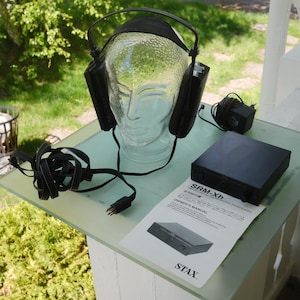 Könnte beinhalten: Schwarzer Kopfhörer auf einem Glaskopf, mit einem schwarzen Verstärker und einer Bedienungsanleitung. Auf dem Verstärker steht "SRM-Xh". Ein Netzkabel und ein schwarzes Netzteil sind ebenfalls vorhanden.