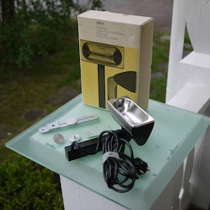May include: A vintage Braun film projector lamp with its original box and accessories. The lamp has a silver reflector and a black base. The box is beige and yellow with the Braun logo and product information. Additional parts include a metal bracket and two small silver discs.