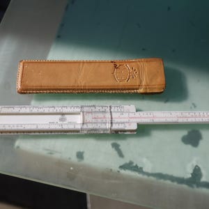 May include: A white slide rule with red and black markings, next to a tan leather case. The slide rule has a sliding center section and a projecting arm. The case has a decorative embossed design.