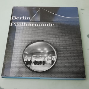 May include: A book cover featuring the words "Berlin/Philharmonie" in white text. The cover has a black and white photograph of a building with a circular window showing people inside. The top of the cover is blue.