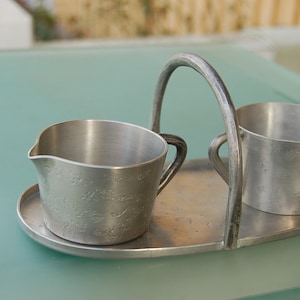 May include: A vintage silver creamer and sugar bowl set. The set includes a creamer with a spout, a sugar bowl, and a tray with a handle. The metal has a textured surface and a classic design, perfect for serving coffee or tea.