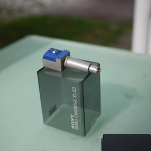 May include: A silver and blue Sony Stereo Cartridge XL-33 stylus with a clear plastic case. The stylus has a silver body and a blue top. The case is a dark gray color. The text "SONY STEREO CARTRIDGE XL-33" is printed on the case.