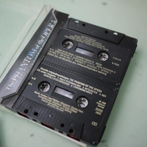 May include: A black cassette tape case with two cassettes inside. The case has the title "The Phantom of the Opera" printed on it. The cassettes are black with white text, and the labels list song titles and credits.