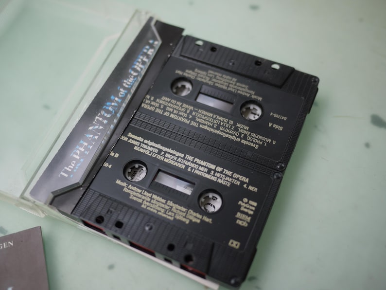 May include: A black cassette tape case with two cassettes inside. The case has the title "The Phantom of the Opera" printed on it. The cassettes are black with white text, and the labels list song titles and credits.