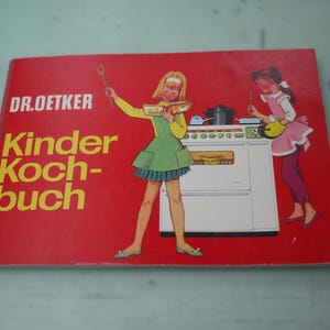 May include: A red Dr. Oetker Kinder Kochbuch cookbook featuring a graphic illustration of two individuals in aprons cooking at a white stove. The text on the cover is printed in white and yellow, showing the brand name and the title of the book.
