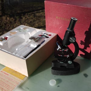 May include: A black microscope with a red box labeled "Microscope". The microscope is next to an open box with various scientific tools and slides. A small white cap is in front of the microscope.