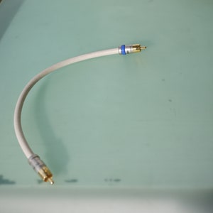 Puede incluir: Cable de audio blanco con conectores dorados en ambos extremos. El cable tiene una banda azul y plateada cerca de un conector. El cable está curvado y descansa sobre una superficie verde claro.