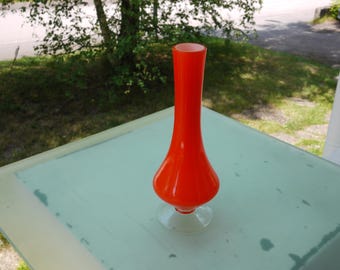 May include: A vibrant orange glass vase with a clear, circular base. The vase has a bulbous lower section that tapers to a slender neck and a flared opening. The vase is displayed on a light green surface, with a natural outdoor background.