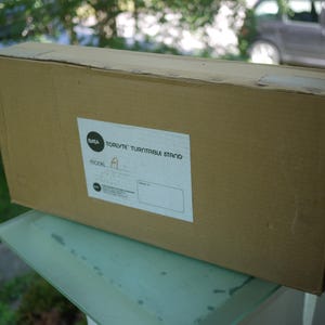 May include: A rectangular brown cardboard shipping box with a white label on the front. The label features the text "RATA TORLYTE TURNTABLE STAND" and "MODEL A" written in black ink. The box is sealed with clear adhesive tape along the top edges.