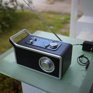 Könnte beinhalten: Ein schwarzes und silbernes tragbares Radio mit einem silbernen Griff, einer Antenne und Bedienknöpfen. Das Radio steht auf einer Glasoberfläche mit einem Netzteil und Kabel. Das Radio hat ein Zifferblatt und einen Lautsprecher.