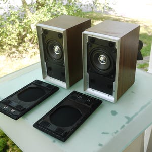 Könnte beinhalten: Zwei braune und silberne Lautsprecher mit schwarzen Lautsprecherabdeckungen. Die Lautsprecher haben eine silberne Umrandung und einen schwarzen Konus. Zwei schwarze Lautsprecherabdeckungen liegen vor den Lautsprechern.