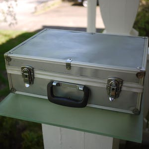 May include: A silver-colored aluminum briefcase with a black handle and two latches. The case is rectangular with rounded corners and sits on a clear glass shelf. The background is blurred, suggesting an outdoor setting.