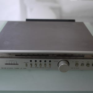 以下が含まれることがあります： シルバーとグレーのSABA FM/AMステレオチューナーCT 300。前面にはチューニングダイヤル、ボタン、大きなノブがあります。上部はグレーで、ユニットはガラスの表面にあります。「SABA FM/AM STEREO TUNER CT 300」の文字が見えます。