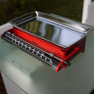 May include: A vintage red kitchen scale with a stainless steel tray. The scale has a black and white measurement indicator, with a maximum capacity of 12 kg. The scale is a classic design, ideal for measuring ingredients.