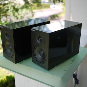 Könnte beinhalten: Zwei schwarze Yamaha-Lautsprecher mit glänzendem Finish werden auf einer Glasoberfläche präsentiert. Jeder Lautsprecher hat einen runden Tieftöner und einen kleineren Hochtöner. Die Lautsprecher sind rechteckig und scheinen neu zu sein.