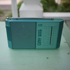 Könnte beinhalten: Ein türkisfarbenes ITT Tiny 505 Kofferradio mit rechteckigem Gehäuse, einer silbernen Teleskopantenne an der Oberseite, einem Lautsprechergitter auf der linken Seite und einer Frequenzanzeige rechts mit dem Aufdruck ITT und tiny 505.