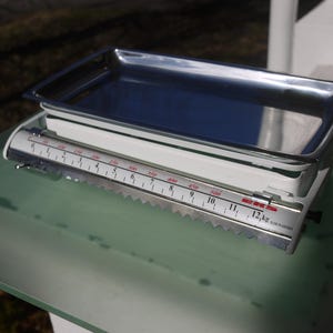 May include: A vintage white and silver kitchen scale with a stainless steel tray. The scale has a measurement indicator with numbers and a maximum weight of 12 kg. The scale is on a green surface.