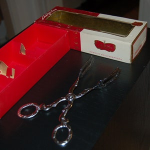 May include: A red and white box with text and a silver serving utensil. The box is open, revealing a gold interior. The serving utensil is ornate with a decorative handle. The box has the text "Chokladfabriken".