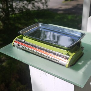 May include: A vintage-style mechanical kitchen scale with a lime green metal base and a removable rectangular stainless steel tray. The front display features a horizontal orange scale with black numerical markings and the brand name Terra.
