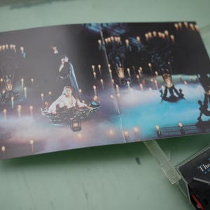 May include: An open cassette case insert featuring a scene from "The Phantom of the Opera." The image shows a woman in a boat surrounded by candles, with a man in a cape standing behind her. The cassette case is partially visible.