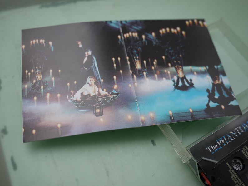 May include: An open cassette case insert featuring a scene from "The Phantom of the Opera." The image shows a woman in a boat surrounded by candles, with a man in a cape standing behind her. The cassette case is partially visible.