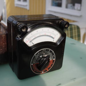 May include: A vintage black electrical meter with a white gauge and red markings. The meter has a circular dial and two knobs. A brown leather case is visible in the background. The meter is likely used for measuring electrical current or voltage.
