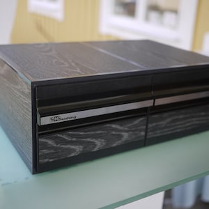 May include: A black wooden box with a textured grain pattern. The box has a rectangular shape with two drawers and a top section. The brand name "SH Sunhing" is visible on a silver label on the front.