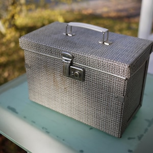 May include: A vintage grey and white patterned box with a silver handle and clasp. The box is rectangular and appears to be made of a woven fabric. The handle and clasp are made of metal.