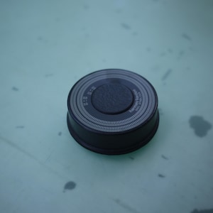 Op de afbeelding: Een ronde zwarte Harman Kardon luidspreker met een textuur in het midden en een omliggende ring met een fijn wit stippenpatroon. Het apparaat heeft een lage cilindrische vorm met een matte afwerking en duidelijke merktekst op de bovenkant.