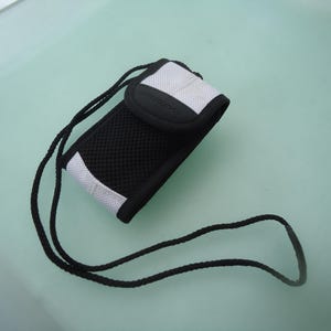 May include: Black and white cell phone case with a mesh front and a black cord. The case has white accents and a black flap. The cord is black and appears to be made of a woven material. The case is designed to hold a cell phone.