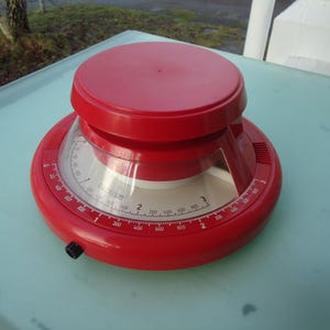 May include: A red kitchen scale with a circular design. The scale has a clear, curved window displaying a measurement dial with numbers and markings. The top and base are solid red, with a black knob on the side. The scale is designed for measuring ingredients.
