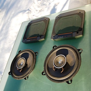 Puede incluir: Cuatro altavoces de audio para coche y sus cubiertas están dispuestos sobre una superficie verde claro. Los altavoces son negros con un cono central y cableado de cobre. Las cubiertas son negras con una pantalla de malla.