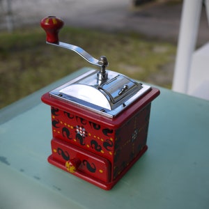 Puede incluir: Un molinillo de café manual rojo y plateado con manivela. El cuerpo de madera está pintado con diseños florales y de volutas en blanco y negro. El molinillo tiene un pequeño cajón y una tapa plateada.