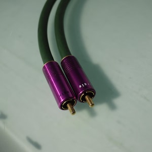 Könnte beinhalten: Zwei Audiokabel mit lila Steckern und goldfarbenen Spitzen. Auf den Steckern steht das Wort "qig". Die Kabel sind dunkelgrün und gebogen. Der Hintergrund ist hell und einfarbig.