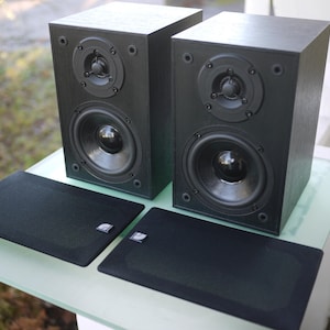 Puede incluir: Dos altavoces de madera negros con un tweeter y un woofer, y dos cubiertas de altavoz negras. Los altavoces son rectangulares y se encuentran sobre una superficie de vidrio. Las cubiertas de los altavoces tienen un pequeño logotipo en la esquina.
