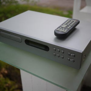 Könnte beinhalten: Ein silberner, rechteckiger elektronischer Mediaplayer mit einem Frontlader-Disc-Fach, einem zentralen Anzeigefenster und einem Bedienfeld mit mehreren runden Tasten. Eine schwarze Fernbedienung mit zahlreichen kleinen Tasten liegt oben auf.