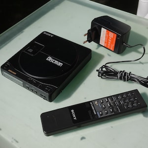 Könnte beinhalten: Ein schwarzer tragbarer Sony Discman CD-Player mit Fernbedienung und Netzteil. Auf dem Discman steht "Discman". Die Fernbedienung hat mehrere Tasten. Das Netzteil ist schwarz mit einem orangefarbenen Etikett.