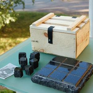 May include: A wooden crate with a black leather strap and a lid sits on a glass surface. Several black, square-shaped stones and a rectangular stone tile with a grid pattern are arranged nearby. A small printed image is also present.