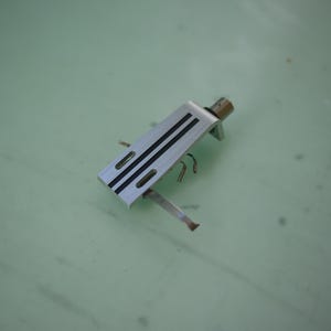May include: A silver-toned metal turntable headshell featuring two parallel black stripes running along the top surface, two elongated rectangular slots, and a small metal connector pin extending from the rear end of the component.