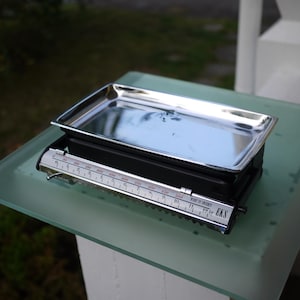 May include: A vintage black and silver scale with a stainless steel tray. The scale has a measurement bar with numbers and markings. The scale is sitting on a glass surface.