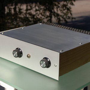 May include: A silver-colored electronic device with two black knobs and a small button. The top is a brushed metal finish, and the sides are wood-toned. The device sits on a glass surface, suggesting a component for audio equipment.