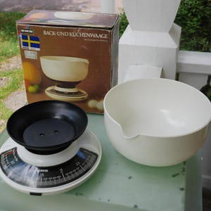 May include: A vintage kitchen scale with a black bowl and a white base, alongside a large white mixing bowl. The scale has a circular dial with measurements in grams. The box reads "BACK-UND KÜCHENWAAAGE".
