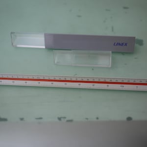 May include: A white and red ruler with centimeter and millimeter markings, a clear plastic case, and a gray case with the text "LINEX". The items are on a light green surface.