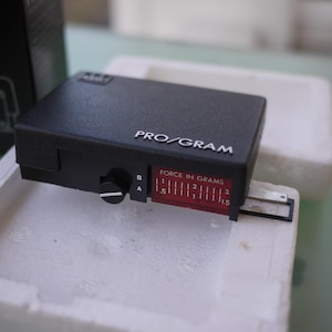 Könnte beinhalten: Ein schwarzes Pro/Gram-Tonabnehmer-Ausrichtungswerkzeug mit einer rot-weißen Kraftanzeige, gemessen in Gramm. Das Werkzeug liegt auf einem weißen Schaumstoffeinsatz, im Hintergrund ist eine schwarze Box zu sehen.