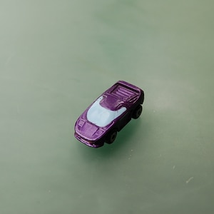 Puede incluir: Un pequeño coche de juguete morado con un parabrisas azul claro. El vehículo en miniatura tiene ruedas negras y está sobre un fondo verde claro. El coche de juguete es una réplica en miniatura de un coche deportivo.