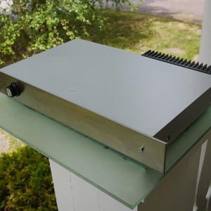Puede incluir: Un dispositivo electrónico plateado con una perilla negra y un componente negro y estriado en la parte superior. El dispositivo rectangular se encuentra sobre una superficie de vidrio, con vegetación en el fondo.